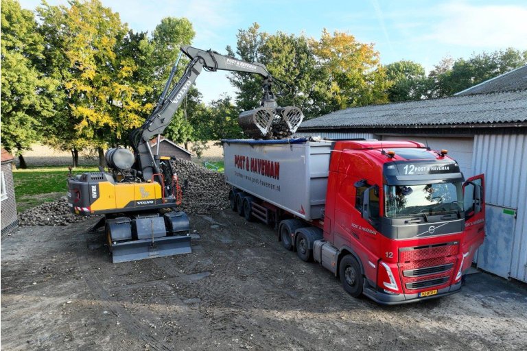 Een kraan die een vrachtwagen van Post & Haveman laadt met bieten. De vrachtwagen staat voor een schuur, de bieten worden vanaf de grond geladen.